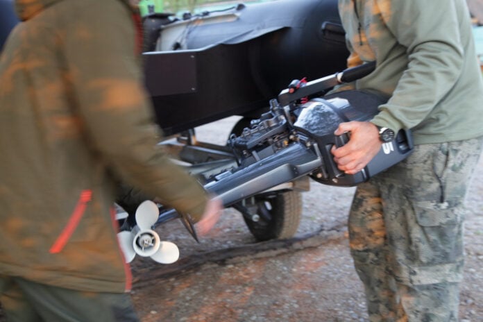 Two,Men,Carry,An,Outboard,Motor,Against,The,Background,Of